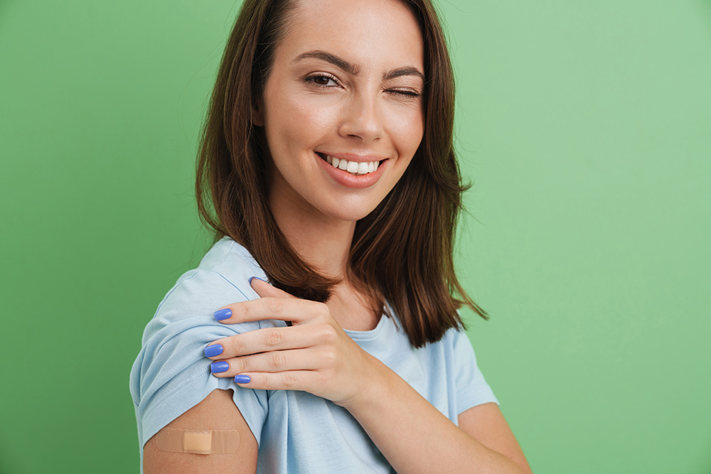 Nainen vinkkaa silmää kameralle ja nostaa hihaa näyttäen rokotelaastarin kädessään